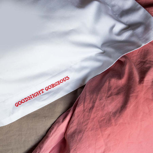 A close-up of a white pillowcase with red embroidered text reading 'GOODNIGHT GORGEOUS', layered over a dusty pink sheet and a beige duvet.