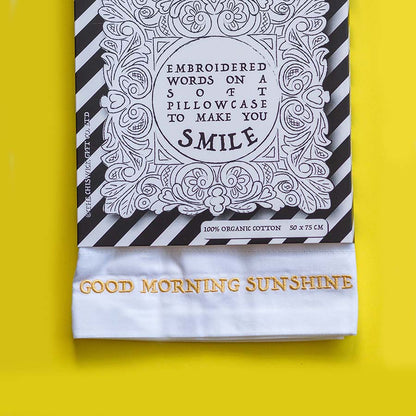 A folded white pillowcase with gold embroidered text reading 'GOOD MORNING SUNSHINE' is displayed in front of its black-and-white striped packaging card on a bright yellow background.