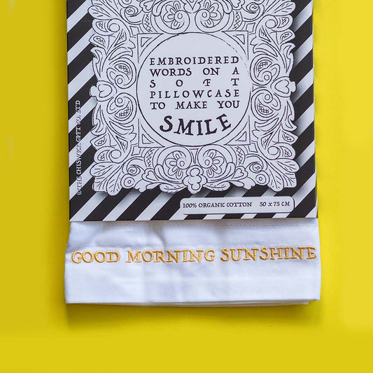 A folded white pillowcase with gold embroidered text reading 'GOOD MORNING SUNSHINE' is displayed in front of its black-and-white striped packaging card on a bright yellow background.